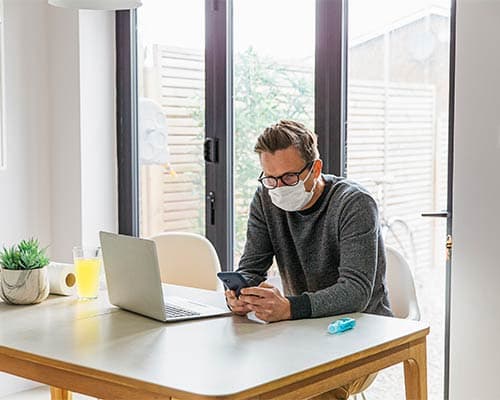 A worker sitting in his home office with a mask on and hand sanitiser and toilet roll on the table